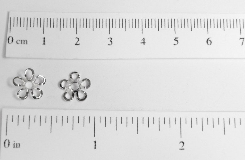 May include: Two small, silver-colored flower-shaped beads. The beads are next to a ruler showing inches and centimeters. Each bead has five petals and a central hole. The beads are likely used for jewelry making or crafting.