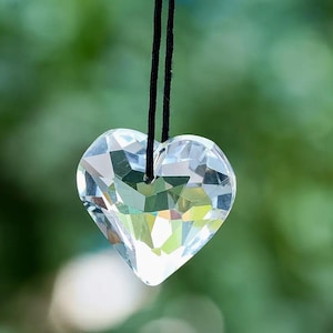 May include: A clear, faceted crystal heart pendant hanging from a black cord. The heart is cut with numerous facets, reflecting light and creating a sparkling effect. The background is a soft, blurred green.