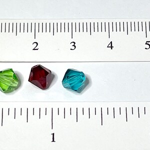 May include: Four faceted glass beads in different colors: purple, green, red, and blue. The beads are approximately 1 cm in diameter and are shown next to a ruler.