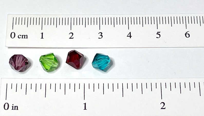 May include: Four faceted glass beads in different colors: purple, green, red, and blue. The beads are approximately 1 cm in diameter and are shown next to a ruler.