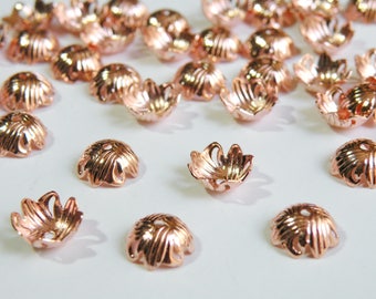 May include: A group of rose gold metal bead caps with a decorative scalloped design. The caps are scattered on a white surface.