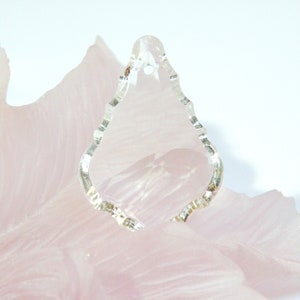 以下が含まれることがあります： ピンクの表面に置かれた、波打った縁のある透明なクリスタル製の涙型のペンダント。ペンダントは紐から吊り下げられています。