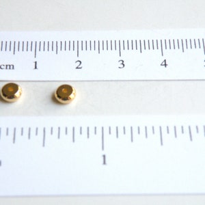 May include: Two gold-colored metal beads, each with a hole in the center, are shown on a white surface next to a ruler. The ruler is marked in inches and centimeters.