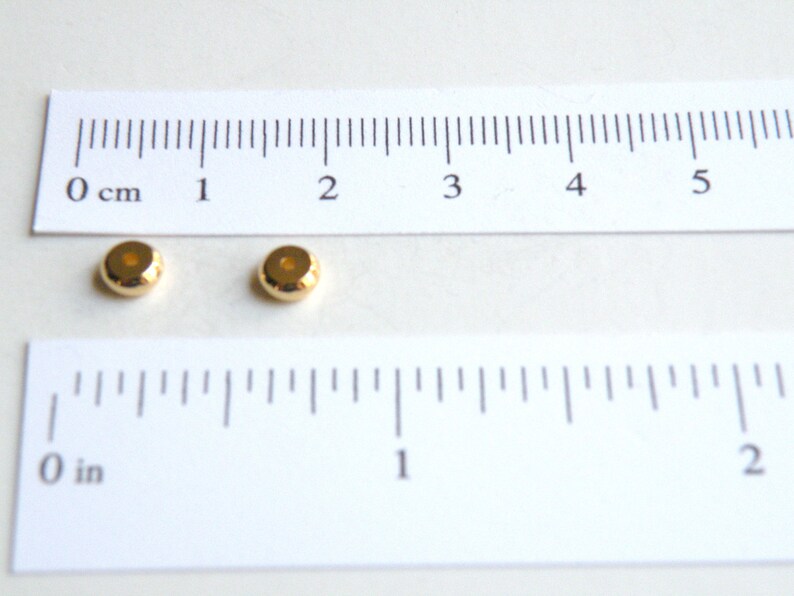 May include: Two gold-colored metal beads, each with a hole in the center, are shown on a white surface next to a ruler. The ruler is marked in inches and centimeters.