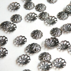 Op de afbeelding: Een verzameling zilverkleurige metalen kralencaps met een decoratief, bloemig ontwerp. De caps zijn verspreid over een wit oppervlak.