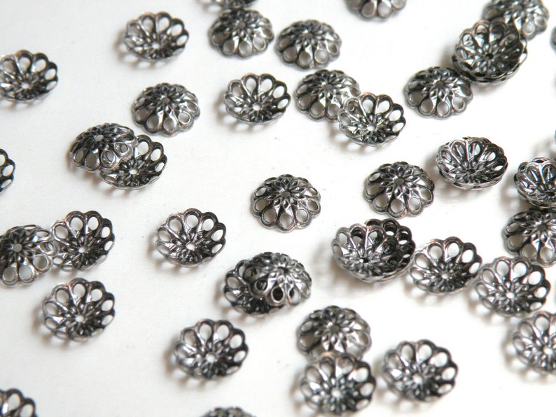 Op de afbeelding: Een verzameling zilverkleurige metalen kralencaps met een decoratief, bloemig ontwerp. De caps zijn verspreid over een wit oppervlak.