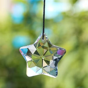 May include: A faceted crystal star pendant hanging from a black string. The star is clear with multiple reflective surfaces, catching light and displaying a rainbow of colors. The background is a soft, blurred green.