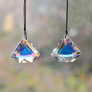 Peut inclure: Deux capteurs solaires en cristal transparent en forme de prisme, suspendus à des cordons noirs. Les cristaux sont facettés, reflétant un arc-en-ciel de couleurs. L'arrière-plan est un mélange flou de verts et de bruns.