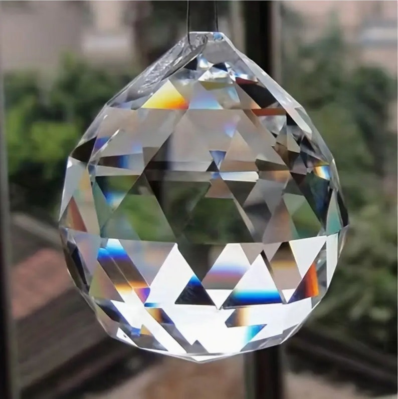 May include: A clear crystal ball with a faceted design. The ball is hanging from a silver string and is reflecting rainbow colors.