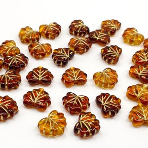 May include: A collection of amber glass leaf-shaped beads with gold accents. The beads are scattered on a white surface.