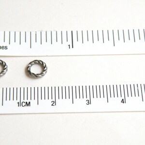 K&ouml;nnte beinhalten: Zwei silberne Sprungringe, jeder mit einem Durchmesser von etwa 1,2 cm, werden neben einem Lineal gezeigt. Die Sprungringe sind verdreht und haben eine strukturierte Oberfl&auml;che.
