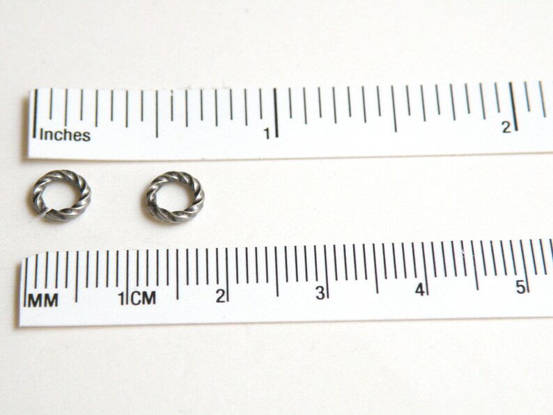 K&ouml;nnte beinhalten: Zwei silberne Sprungringe, jeder mit einem Durchmesser von etwa 1,2 cm, werden neben einem Lineal gezeigt. Die Sprungringe sind verdreht und haben eine strukturierte Oberfl&auml;che.