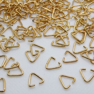 May include: A scattering of small, gold-coloured triangle-shaped jewellery findings. Each finding has a triangular shape with an open side, and they are scattered across a white surface. These findings are likely used for jewellery making.