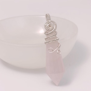 May include: A pale pink, faceted, pointed crystal pendant wrapped in silver-coloured wire. The wire forms a spiral design at the top and secures the stone with decorative loops. The pendant has a smooth, polished surface and a tapered, geometric shape.