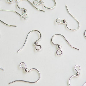 May include: Close-up of several silver-colored earring hooks, each with a curved wire and a small ball at the end. The hooks are scattered on a white surface, showcasing their simple, elegant design. These are ideal for DIY jewelry making.
