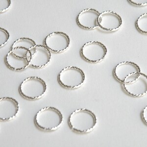 May include: A collection of silver jump rings, each with a twisted design. The rings are arranged in a random pattern on a white background.