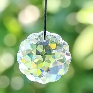 May include: A clear, faceted crystal suncatcher with a scalloped edge, suspended by a black string. The crystal reflects a spectrum of colors, with a blurred green background.