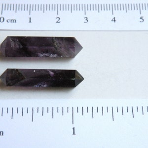 May include: Two dark purple amethyst points, each approximately 5 cm long, are displayed on a white surface with a ruler for scale.