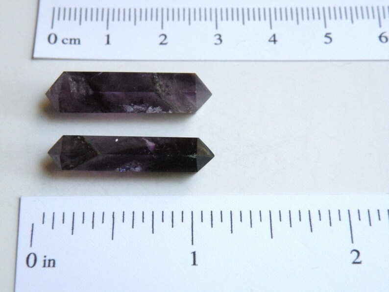 May include: Two dark purple amethyst points, each approximately 5 cm long, are displayed on a white surface with a ruler for scale.