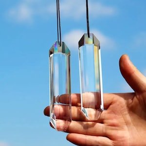 May include: Two clear crystal prisms hanging from black cords. The prisms are faceted and have a rectangular shape. The prisms are held in a hand against a blue sky background.