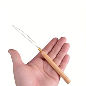 May include: A hair extension tool with a light wooden handle and a thin metal loop. The handle has a gold-colored metal accent at the top. The tool is designed for applying hair extensions.