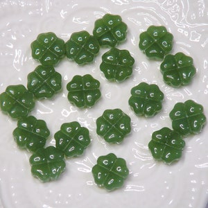 May include: A collection of 17 green four-leaf clover shaped glass beads. The beads are arranged on a white surface with a decorative pattern.