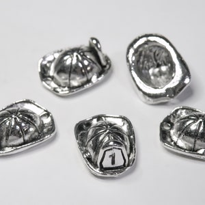 May include: Five silver-colored miniature firefighter helmet charms. Each charm has a detailed design, including a brim, a top with ridges, and a small handle. One charm has the number "1" on the front.