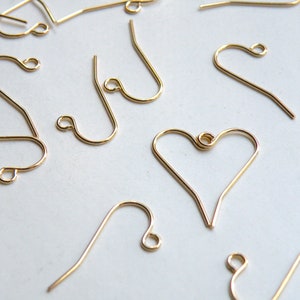May include: A close-up of a group of gold-colored earring hooks. Some of the hooks are shaped like a heart, while others are a simple curved shape.