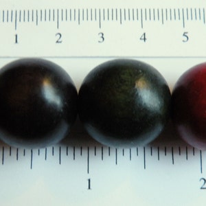 May include: Close-up of four round, dark-colored beads. The beads are arranged in a row next to a ruler marked in inches and centimeters. The beads range in color from dark purple to red.