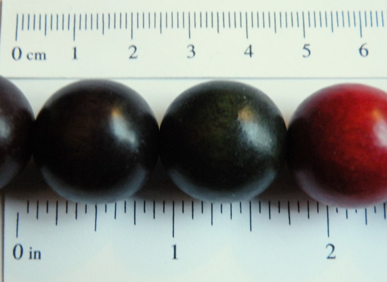 May include: Close-up of four round, dark-colored beads. The beads are arranged in a row next to a ruler marked in inches and centimeters. The beads range in color from dark purple to red.