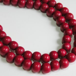 May include: A close-up of a necklace featuring round, deep red wooden beads. The beads are strung together, creating a curved pattern against a white background. The necklace is a simple, yet elegant accessory.