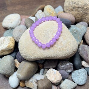 May include: A light purple beaded bracelet rests on a light-colored stone, surrounded by various smooth rocks. The bracelet is made of small, round beads. The background is a wooden surface.