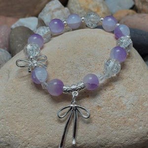 May include: A beaded bracelet featuring alternating purple and clear crystal beads. The bracelet has silver-toned bow-shaped accents and a dangling charm. The bracelet is displayed on a light-colored stone, with other stones in the background.