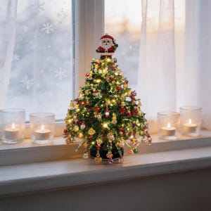 Op de afbeelding: Een miniatuur kerstboom gemaakt van groene kralen, versierd met kleine kleurrijke hangende ornamenten en warmwitte kerstverlichting. Een figuurtje van de Kerstman van kralen zit boven op de boom, die op een ronde zwarte voet staat.