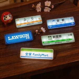 Könnte beinhalten: Eine Sammlung beleuchteter japanischer Schilder und eine Daruma-Puppe auf einer Holzoberfläche. Die Schilder zeigen japanischen Text und englische Übersetzungen, darunter "Fuji", "Kyoto", "Akihabara" und "FamilyMart". Die rechteckigen Schilder haben einen Leuchteffekt.