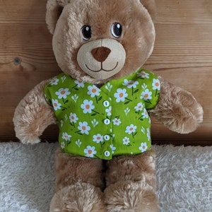 May include: A brown teddy bear wearing a green shirt decorated with white daisy flowers. The shirt has white buttons. The bear has a smiling face with black eyes and a brown nose. The bear is on a white fluffy surface.