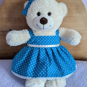 Può includere: Un orsacchiotto di peluche color crema con un vestito blu a pois bianchi e un fiocco abbinato. L'abito presenta pois bianchi e un bordo in pizzo bianco. L'orso ha un naso marrone e occhi neri ed è in posa con le braccia tese.