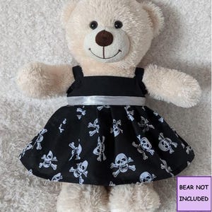 May include: A beige plush teddy bear wearing a black sleeveless dress with a white skull and crossbones pattern. The dress features a solid black bodice and a silver ribbon waistband. A purple box with the text BEAR NOT INCLUDED is visible.