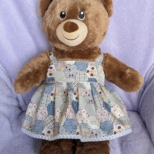 May include: A brown teddy bear wearing a patchwork dress with a floral and geometric pattern. The dress has a square neckline and white lace trim. The bear has a light brown snout and a smiling expression.
