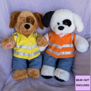 May include: Two plush dog toys wearing blue denim trousers and high-visibility safety vests. The left dog is brown with a yellow vest, and the right dog is white with a black ear patch and an orange vest. A purple box states BEAR NOT INCLUDED.