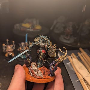 Puede incluir: Una figura en miniatura detallada de un guerrero con armadura azul y dorada, sosteniendo una espada y un escudo brillantes. La figura se encuentra sobre una base circular con un esqueleto y otras figuras. La miniatura se sostiene en una mano.