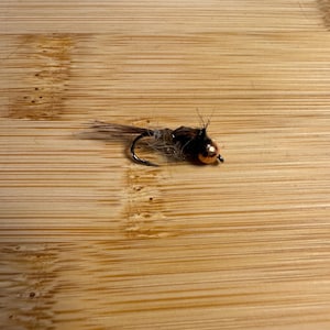 Può includere: Primo piano di una mosca da pesca con un amo nero, una perlina color rame e piume marroni e beige. La mosca è progettata per imitare un insetto e viene utilizzata per attirare i pesci. La mosca è appoggiata su una superficie di legno.