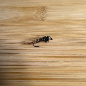 May include: A small, detailed fishing fly with a silver head, dark body, and brown tail fibers. The fly is positioned on a light brown wooden surface, showcasing its intricate design and craftsmanship. This fly is designed for angling.