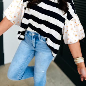 May include: A black and white striped top with floral print sleeves is paired with light blue jeans. The top has a round neckline and a loose fit. The jeans are a light wash with a slim fit.