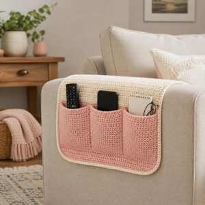 May include: A crocheted sofa armrest organizer in cream and pink. The organizer has three pockets for a remote control, phone, notebook, and glasses. The armrest cover is placed on a beige sofa.