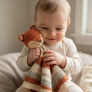 Pode incluir: Um cobertor de segurança para bebé em forma de raposa, feito à mão em tons de castanho, bege e verde. A raposa tem um rosto detalhado com pequenos olhos pretos e um nariz pontudo. O cobertor apresenta padrões listrados e é segurado por um bebé.