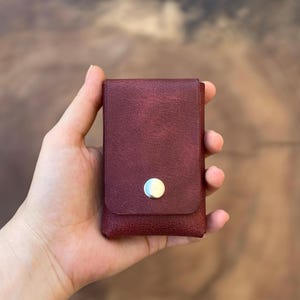May include: A rectangular, deep burgundy wallet with a textured surface and a single silver-toned metal snap button closure on the front flap. The wallet features a vertical orientation and a simple, clean design with visible stitching along the edges.