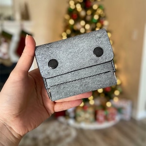 Puede incluir: Una cartera rectangular de fieltro gris con una solapa plegable asegurada por dos botones de presión circulares negros. La cartera tiene costuras visibles a lo largo del borde inferior de la solapa y un diseño compacto y estructurado de tela.
