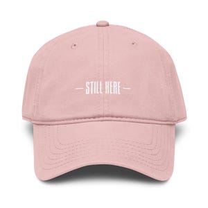 May include: A light pink baseball cap with a curved brim. The cap features the white text "- STILL HERE -" centered on the front. The cap is made of a soft fabric and has a classic six-panel design.