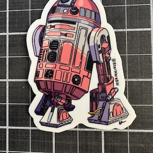 Puede incluir: Una pegatina con una ilustración estilizada de un droide rosa y blanco con detalles morados y negros. El droide tiene una cabeza redondeada y un cuerpo rectangular. El texto "@StarWarsW" es visible.